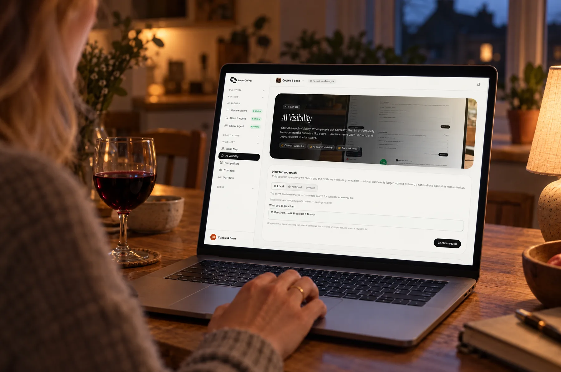 A laptop on a kitchen table in the evening showing Cobble & Bean’s AI visibility results, beside a lamp and a glass of red wine