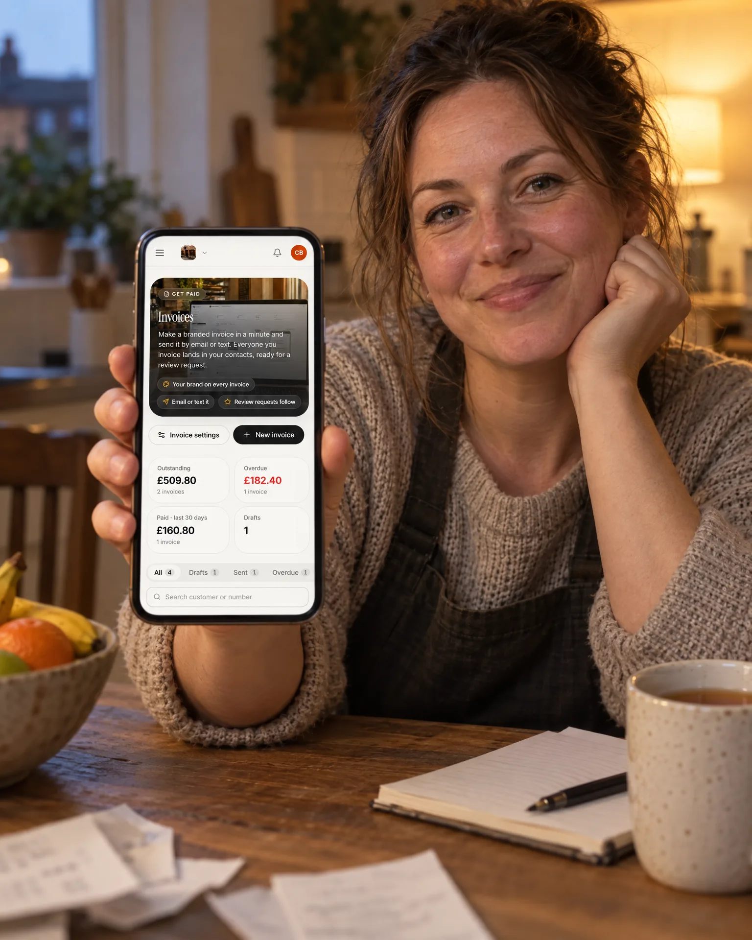 A small business owner at her kitchen table in the evening, showing the Cobble & Bean invoices screen on her phone
