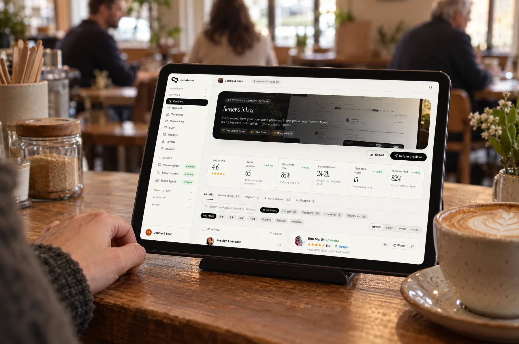 A tablet on the Cobble & Bean counter showing the reviews inbox, next to a fresh cappuccino
