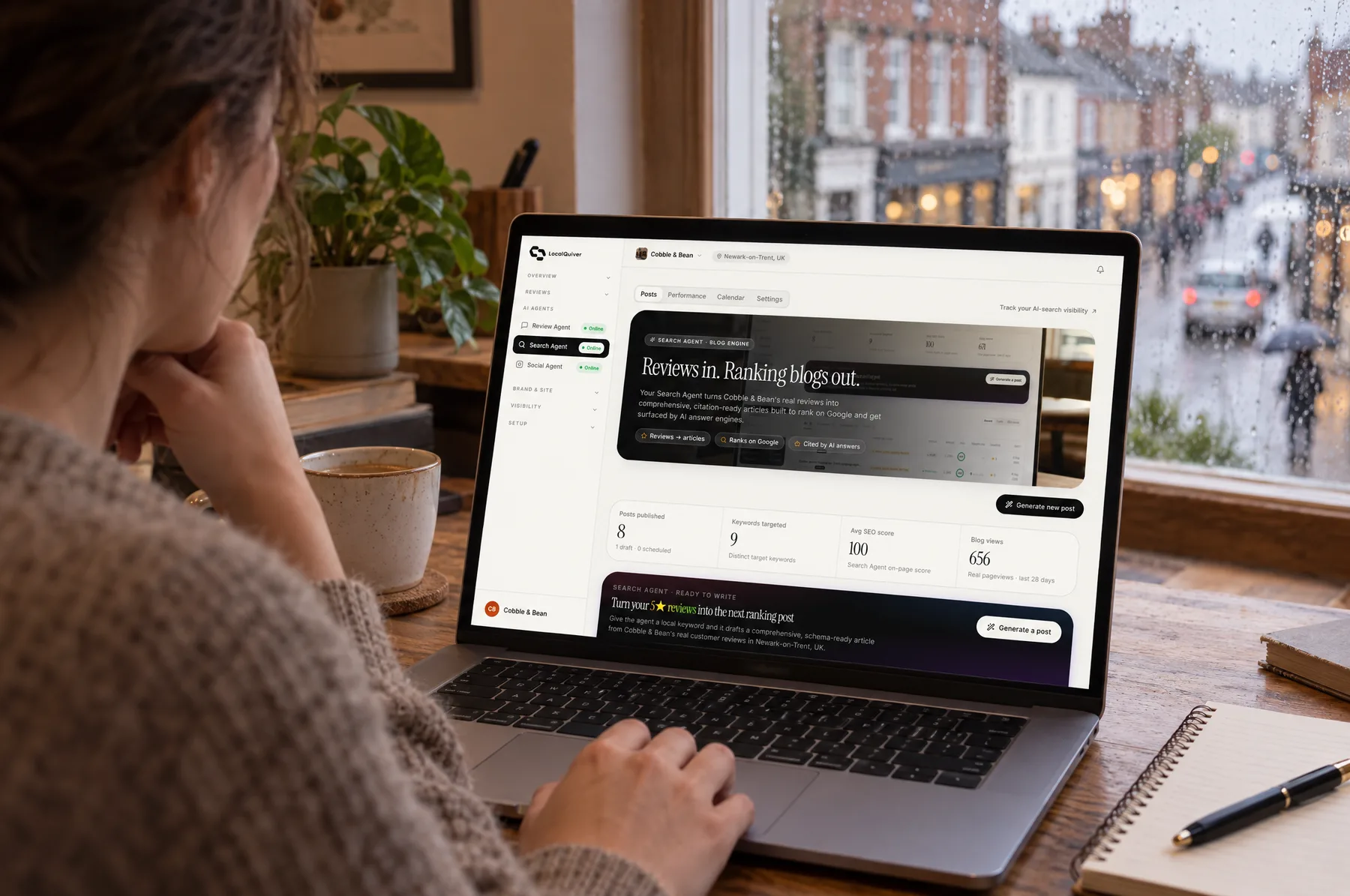 A laptop in a small office above a high street showing Cobble & Bean’s blog and Search Agent dashboard, with rain on the window