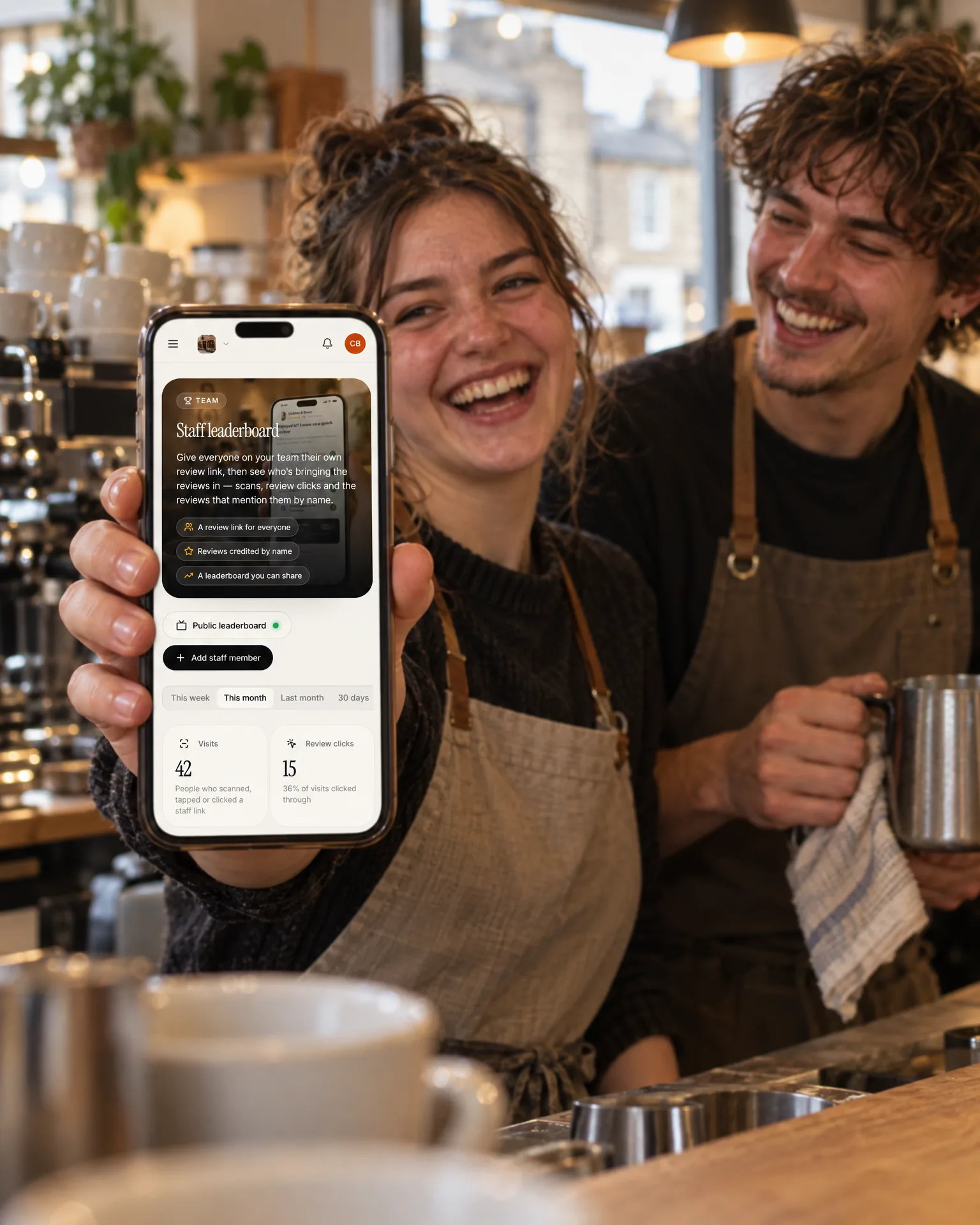 Two Cobble & Bean baristas laughing behind the counter, one holding up a phone showing the café’s staff leaderboard