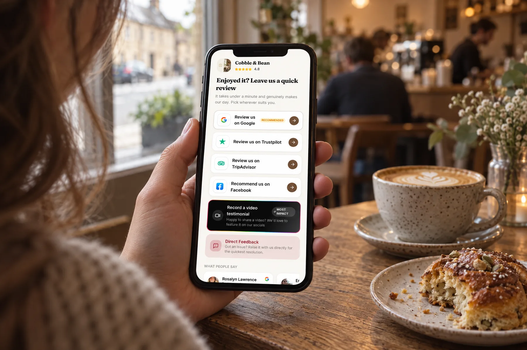 A customer at a Cobble & Bean table holding up their phone on the café’s review page, beside a flat white and a cardamom bun
